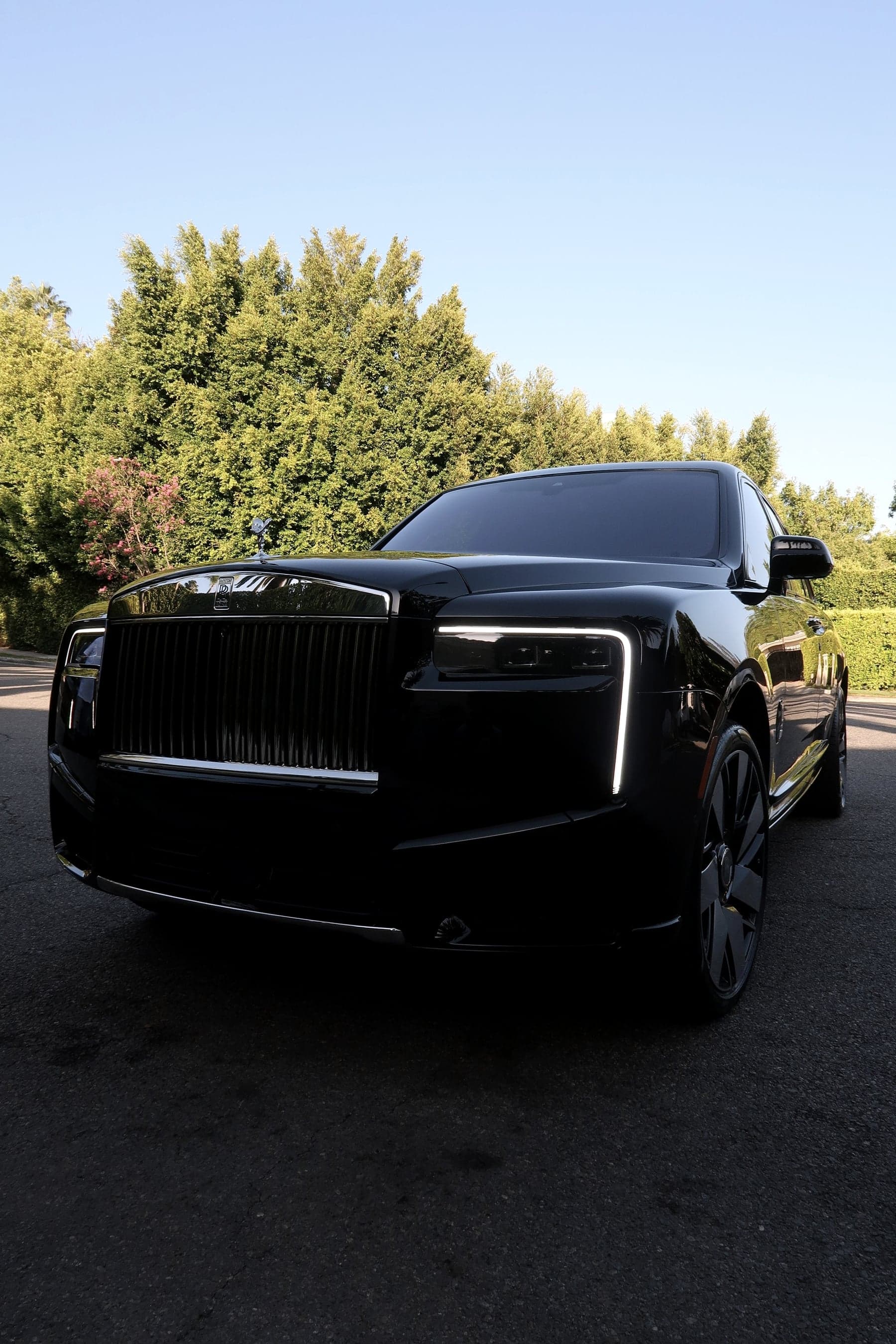 Black 2026 Rolls-Royce Cullinan, low front view with its daytime running lights on, on a tree-lined Los Angeles street