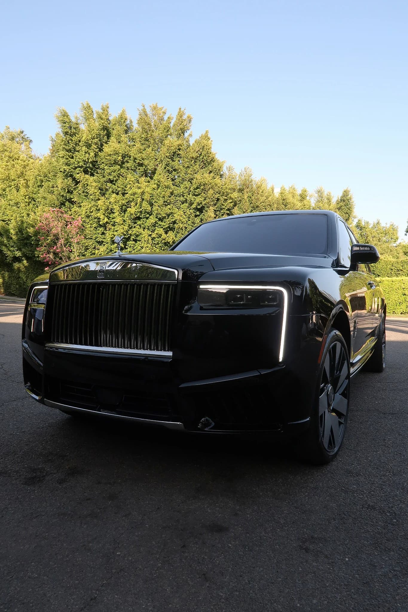 CarPluggLA's black Rolls-Royce Cullinan, low front three-quarter view with its daytime running lights on, parked in front of a tall hedge